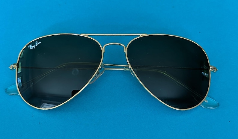 Op de afbeelding: Aviator zonnebril met een goudkleurig montuur en donkere glazen. De zonnebril heeft een dubbele brug en transparante kunststof oortips. Het Ray-Ban logo is zichtbaar op het linkse glas. Op een blauwe achtergrond.