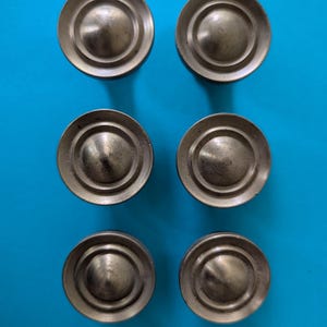 May include: A set of six circular metal cabinet knobs arranged in two columns of three. Each knob features a raised, rounded center dome surrounded by a flat, concentric circular rim with a brushed, antique brass-toned metallic finish.