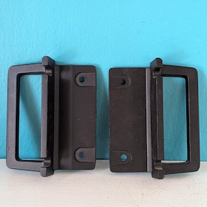 May include: Two black metal cabinet pulls with rectangular handles and mounting plates. Each pull has two screw holes for attachment. The pulls are set against a light blue background.