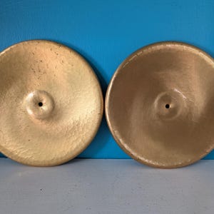 May include: Two circular gold-colored metal discs with a central hole. The left disc features a textured, hammered surface, while the right disc has a smoother, more uniform finish. Both objects are round with slightly raised edges.