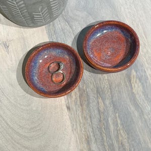 May include: Two small, round ceramic dishes with a reddish-brown and blue glaze. One dish holds a silver ring. The dishes are on a light gray wooden surface, with a gray concrete planter in the background.
