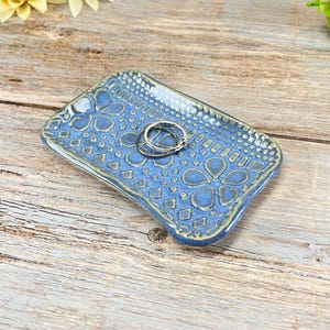 May include: A blue ceramic ring dish with a floral and geometric pattern. The rectangular dish has a raised edge and holds two silver rings. The background includes sunflowers and a wooden surface.