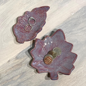May include: Two leaf-shaped ceramic dishes in shades of purple and blue. One dish holds two rings, the other contains several coins. The dishes have a textured surface and are placed on a light gray wooden surface.