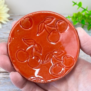 May include: A round, orange ceramic dish with a glossy finish. The dish features an embossed cherry design with leaves. The cherries are evenly spaced across the surface. The dish is held in a hand.
