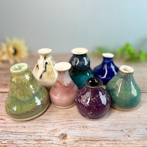 May include: A collection of seven small, handcrafted ceramic vases in various colors. The vases have a bulbous shape with a narrow neck and a glossy finish. Colors include green, white, pink, blue, and purple. The vases are arranged on a wooden surface.
