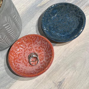 May include: Two small, handmade ceramic ring dishes. One is a vibrant coral color, and the other is a deep blue. Both have a textured, embossed pattern. A pair of rings rests in the coral dish. A gray concrete planter is in the background.