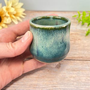 May include: A small, handmade ceramic cup with a teal and blue glaze. The cup has a rounded shape and a slightly textured surface. The rim is a darker green color. The cup is held in a hand, with a wooden surface in the background.