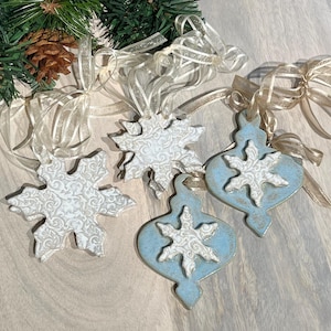 May include: Four ceramic Christmas ornaments. Two are snowflake-shaped, and two are teardrop-shaped with a snowflake design. The ornaments are white and light blue with a textured pattern. Each ornament has a sheer ribbon for hanging.