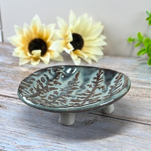 May include: A small, handmade ceramic dish with a teal glaze and a brown tree pattern. The dish is raised on three small, white feet. Decorative sunflowers are in the background.