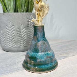 May include: A small, handcrafted ceramic vase with a teal and brown glaze. The vase has a flared base and a narrow neck, holding dried flowers. A potted plant in a gray concrete pot is in the background.
