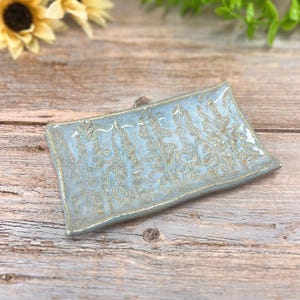 May include: A rectangular ceramic dish with a light blue glaze and a textured, leaf-like pattern. The dish has a slightly raised edge and a subtle, metallic gold accent. The handmade pottery is displayed on a wooden surface.