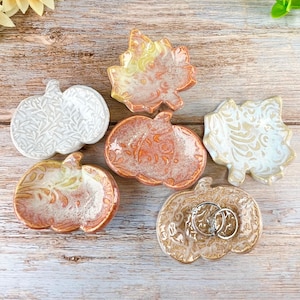 May include: A collection of six small ceramic dishes shaped like pumpkins and leaves, featuring various glazed finishes in white, orange, and tan with intricate embossed patterns. One pumpkin dish contains two silver metal rings.