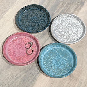 May include: Four small, round ceramic dishes in shades of blue, pink, and white. Each dish features a raised, swirling floral pattern. One pink dish holds two silver rings. The dishes are arranged on a light wood-grain surface.