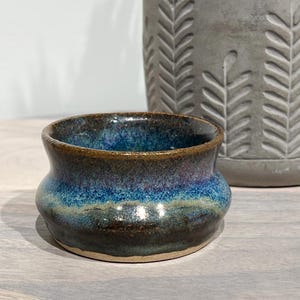May include: A small, handcrafted ceramic bowl with a unique shape and a glossy, multi-toned glaze. The bowl transitions from dark brown at the base to shades of blue and teal, with a brown rim. A gray planter with a leaf pattern is in the background.
