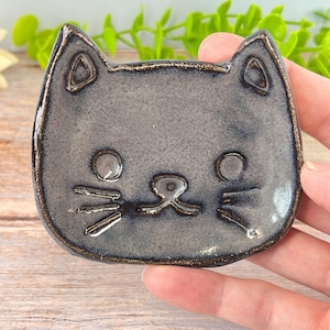 May include: A small, cat-shaped ceramic trinket dish with a speckled grey glaze and dark, metallic-toned edges. The dish features incised details including two pointed ears, circular eyes, a small nose, and three whiskers on each side of the face.