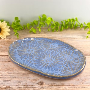 May include: An oval ceramic dish featuring a textured blue glaze with a repeating stamped floral pattern. The edges of the dish are finished with a metallic gold trim, highlighting the irregular, hand-formed shape of the pottery piece.
