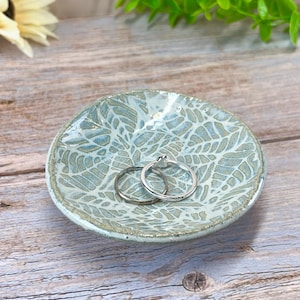 May include: A small, round ceramic dish with a light blue and white leaf pattern. Two silver rings rest inside the dish. The dish is on a wooden surface, with a flower and greenery in the background.