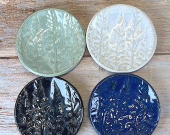 May include: Four circular ceramic dishes featuring an embossed pine tree design. The set includes one sage green, one off-white, one dark navy blue, and one royal blue dish, each with a glossy finish and a slightly raised rim for holding small items.