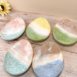 May include: Five handmade ceramic egg-shaped dishes with unique designs. Each dish features a two-tone color scheme with textured patterns. Colors include pink, blue, yellow, green, and brown, with white and gold accents. The dishes are displayed on a wooden surface.