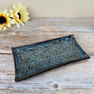 May include: A rectangular ceramic tray with a blue glaze and a detailed brown floral pattern. The tray has a raised edge and is placed on a wooden surface. Two yellow sunflowers are in the background.