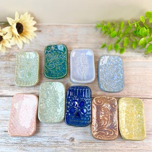 May include: A collection of rectangular ceramic soap dishes in various colors and patterns. The dishes feature embossed designs, including floral and swirling motifs. Colors include blue, green, pink, and yellow. The dishes are arranged on a wooden surface.