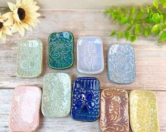 May include: A collection of rectangular ceramic soap dishes in various colors and patterns. The dishes feature embossed designs, including floral and swirling motifs. Colors include blue, green, pink, and yellow. The dishes are arranged on a wooden surface.