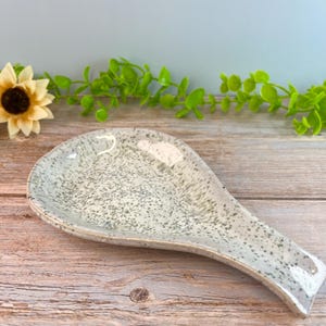 May include: A ceramic spoon rest with a teardrop shape, featuring a light gray base color and a speckled pattern of dark gray dots across the entire surface. The finish is smooth and glossy, reflecting light along the curved edges of the piece.