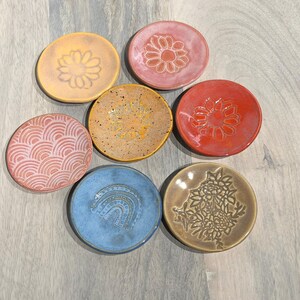 May include: A collection of seven small, round ceramic dishes in various colors, including yellow, pink, red, blue, and brown. Each dish features a unique embossed design, such as flowers, a rainbow, and geometric patterns. The dishes are arranged on a light wood surface.