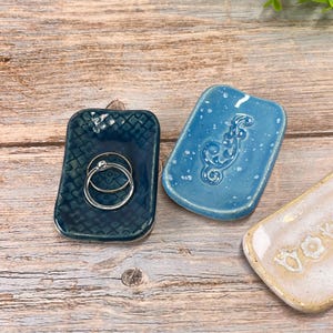 May include: Three rectangular ceramic trinket dishes with rounded corners. One is dark blue with a stamped floral pattern holding two silver rings, one is light blue with a speckled finish and a seahorse relief, and one is cream with text.