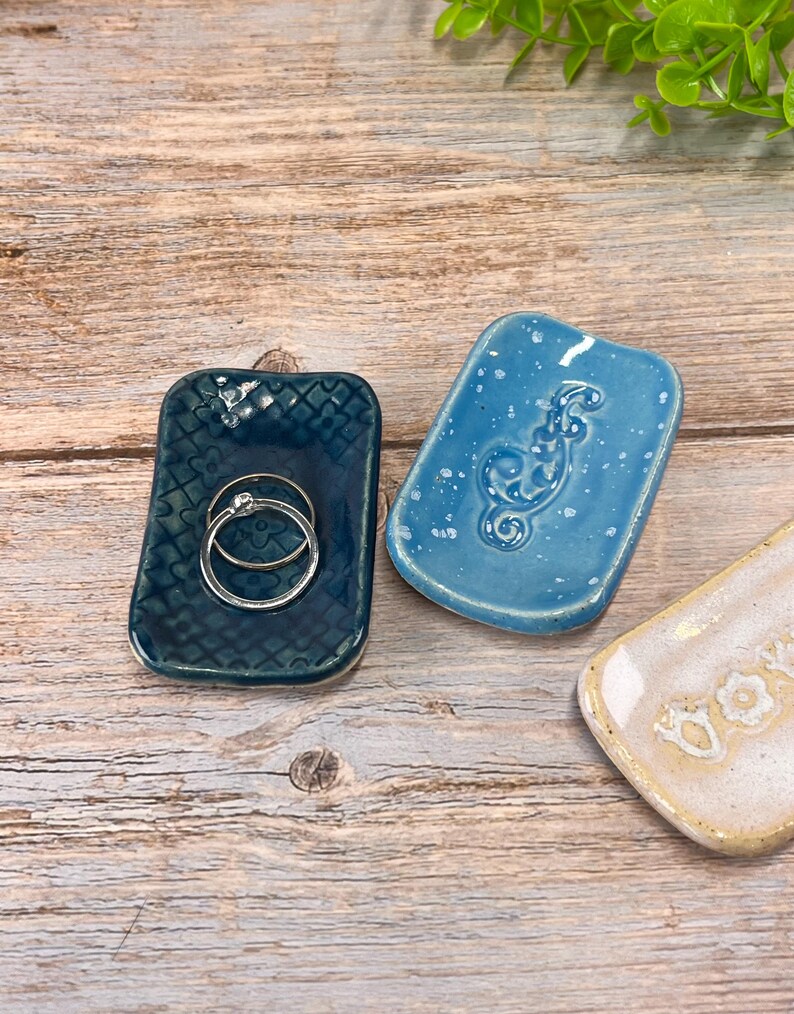May include: Three rectangular ceramic trinket dishes with rounded corners. One is dark blue with a stamped floral pattern holding two silver rings, one is light blue with a speckled finish and a seahorse relief, and one is cream with text.