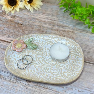 May include: Oval ceramic trinket dish with a cream-colored background and a beige floral pattern. The dish features a pink flower and green leaf accent, two silver rings, and a white tea light candle. The dish is approximately 6 inches long.