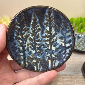 May include: A round, ceramic coaster with a forest design. The coaster features a dark blue background with a textured, tree-like pattern in shades of brown and white. The coaster is held in a hand.