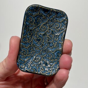 May include: A small rectangular ceramic dish with rounded corners, featuring a glossy glaze in shades of deep blue and muted olive green. The surface is decorated with an intricate, raised relief pattern of swirling, interconnected organic shapes.