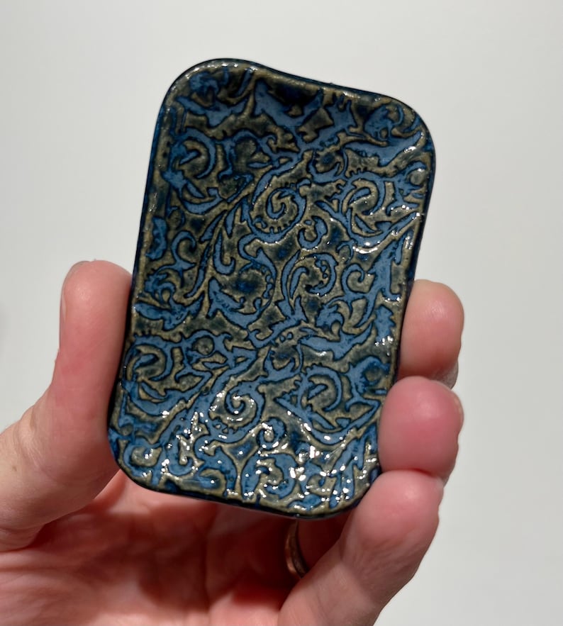 May include: A small rectangular ceramic dish with rounded corners, featuring a glossy glaze in shades of deep blue and muted olive green. The surface is decorated with an intricate, raised relief pattern of swirling, interconnected organic shapes.