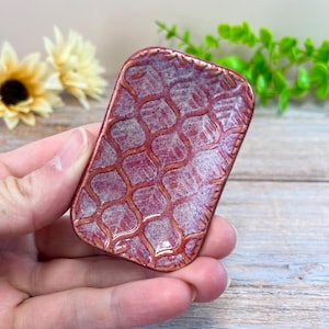 May include: A small rectangular ceramic dish featuring a deep red and purple mottled glaze with a raised, textured lattice pattern across the surface. The edges of the dish are slightly rounded, and the finish has a glossy, reflective appearance.