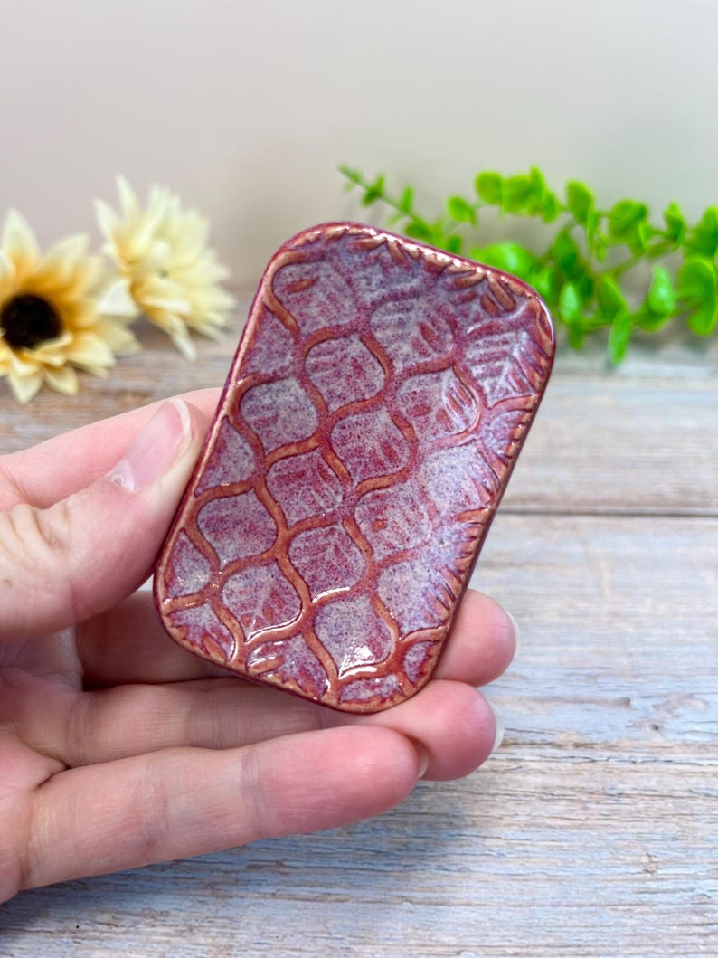 May include: A small rectangular ceramic dish featuring a deep red and purple mottled glaze with a raised, textured lattice pattern across the surface. The edges of the dish are slightly rounded, and the finish has a glossy, reflective appearance.