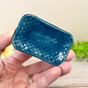 May include: A small, rectangular ceramic dish with a glossy, deep teal glaze. The surface features a raised, repeating lattice pattern with small four-petaled flower shapes at each intersection, creating a textured, grid-like appearance.