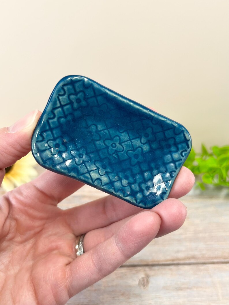 May include: A small, rectangular ceramic dish with a glossy, deep teal glaze. The surface features a raised, repeating lattice pattern with small four-petaled flower shapes at each intersection, creating a textured, grid-like appearance.
