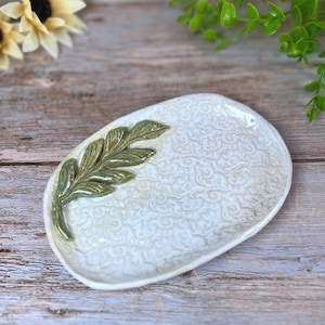 May include: Oval ceramic dish with a white background and a raised, olive green leaf design on the left side. The dish has a textured, swirling pattern. Decorative item.