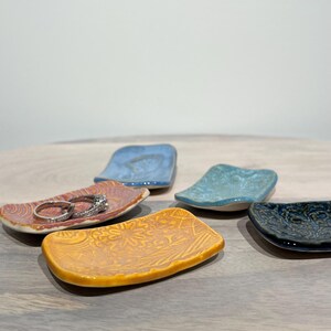 May include: A collection of five small, rectangular ceramic trinket dishes in various colors including yellow, orange, light blue, and dark blue. Each dish features a unique embossed pattern on its surface. Two silver rings rest on the orange dish.