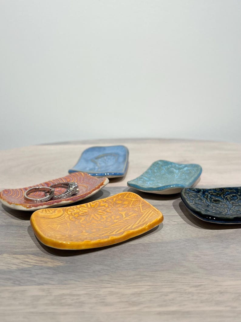 May include: A collection of five small, rectangular ceramic trinket dishes in various colors including yellow, orange, light blue, and dark blue. Each dish features a unique embossed pattern on its surface. Two silver rings rest on the orange dish.