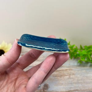 May include: A small, rectangular ceramic dish featuring a deep teal, glossy glaze with a subtle, embossed diamond pattern across the surface. The edges of the dish are slightly raised and have a textured, uneven finish with a light-colored rim.
