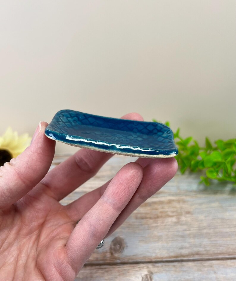 May include: A small, rectangular ceramic dish featuring a deep teal, glossy glaze with a subtle, embossed diamond pattern across the surface. The edges of the dish are slightly raised and have a textured, uneven finish with a light-colored rim.