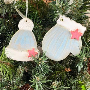 May include: Two ceramic Christmas ornaments in the shape of a hat and a bell. The ornaments are light blue with white trim and a pink star. They hang from a Christmas tree with pine needles and ribbon.