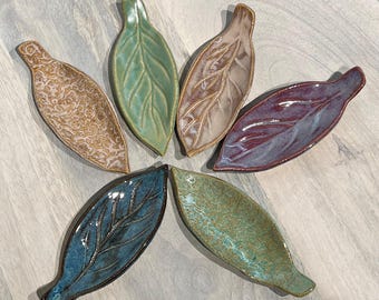 May include: Six ceramic leaf-shaped dishes in various colors, including teal, green, and purple. Some leaves have textured patterns, while others have smooth surfaces. The dishes are arranged in a circular pattern on a light wood surface.