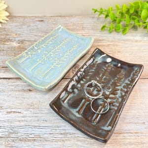May include: Two rectangular ceramic trinket dishes with curved edges, each featuring a debossed pine tree forest design. One dish is light blue, while the other is dark brown with blue highlights. Two silver rings rest on the brown dish.