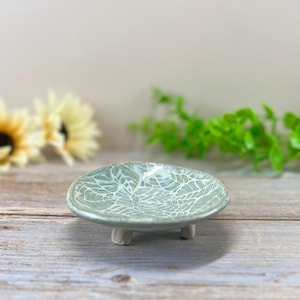 May include: A small, round, ceramic dish with a light blue and white leaf pattern. The dish is elevated on three small, light-colored feet. The background features a wooden surface and blurred greenery.