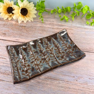 May include: A rectangular ceramic tray with a glossy finish, featuring a dark brown and blue glaze. The surface is embossed with a repeating pattern of vertical fern-like botanical prints that run across the length of the dish.