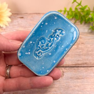 May include: A rectangular ceramic soap dish with a glossy bright blue glaze and a speckled white finish. The center of the dish features a raised, embossed scrollwork pattern. The edges are slightly rounded, and the surface has a smooth, reflective texture throughout the piece.
