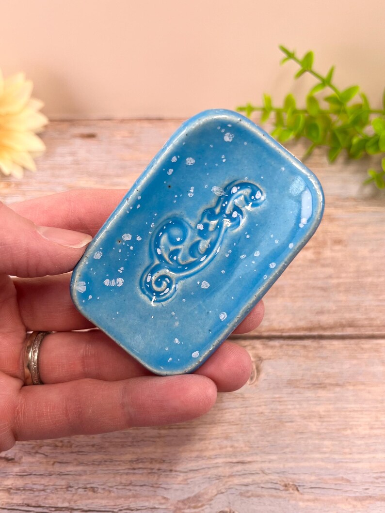 May include: A rectangular ceramic soap dish with a glossy bright blue glaze and a speckled white finish. The center of the dish features a raised, embossed scrollwork pattern. The edges are slightly rounded, and the surface has a smooth, reflective texture throughout the piece.
